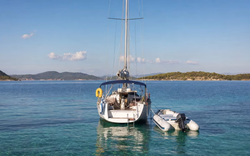 Schlauchboot mit kleinem Außenborder am Heck einer ankernden Segelyacht in einer Bucht