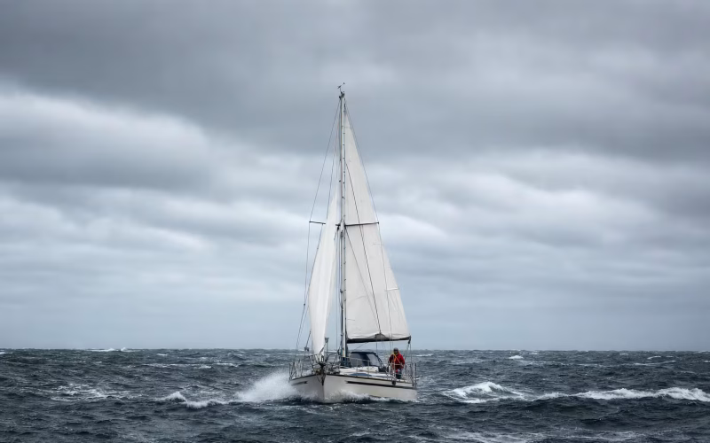 Segelyacht in rauer See mit grauem Himmel, gerefften Segeln und Gischt über dem Bug