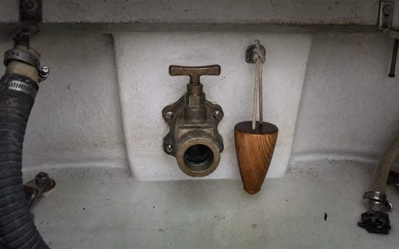 Bronzenes Seeventil im Rumpf mit einem konischen Holzpfropfen an einer Leine daneben zur Leckabwehr