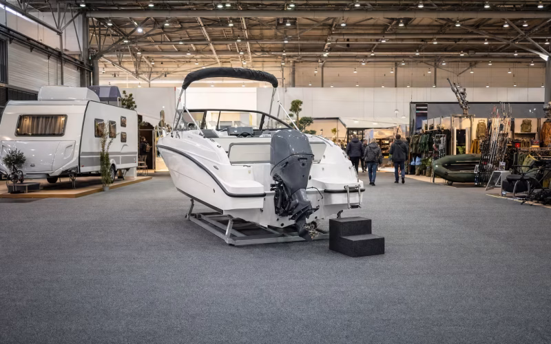 Weißes Trailer-Motorboot mit Außenborder auf einem Messestand, links ein Wohnwagen, rechts Angel- und Outdoorstände in derselben Halle