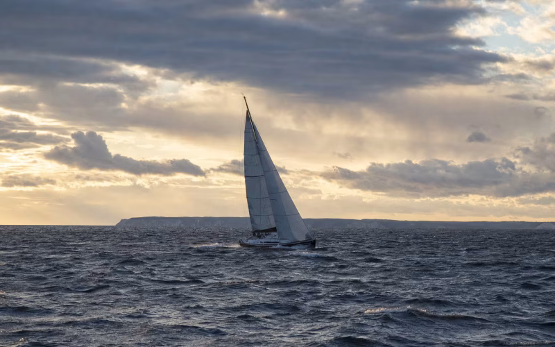 Segelyacht unter Groß und Fock auf bewegter Ostsee im Abendlicht, am Horizont die Steilküste einer Insel